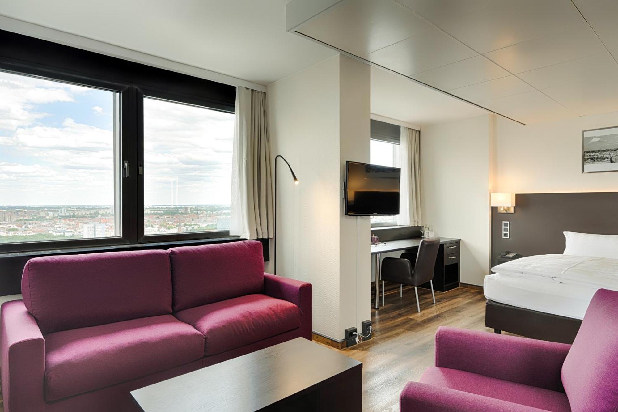 Park Inn By Radisson Berlin Alexanderplatz Esterno foto The photo shows a modern hotel room with a stylish design. It features a seating area with two purple sofas and a coffee table. Opposite the seating area, there is a desk with a chair and a television mounted on the wall. The room has large windows a