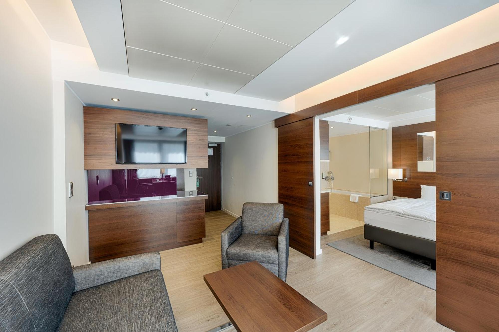 Park Inn By Radisson Berlin Alexanderplatz Esterno foto The photo shows a modern hotel room or suite. On one side, there is a small seating area furnished with a gray sofa and two chairs. A wooden coffee table is positioned in front of the sofa. The wall features a flat-screen TV mounted above a low cabin