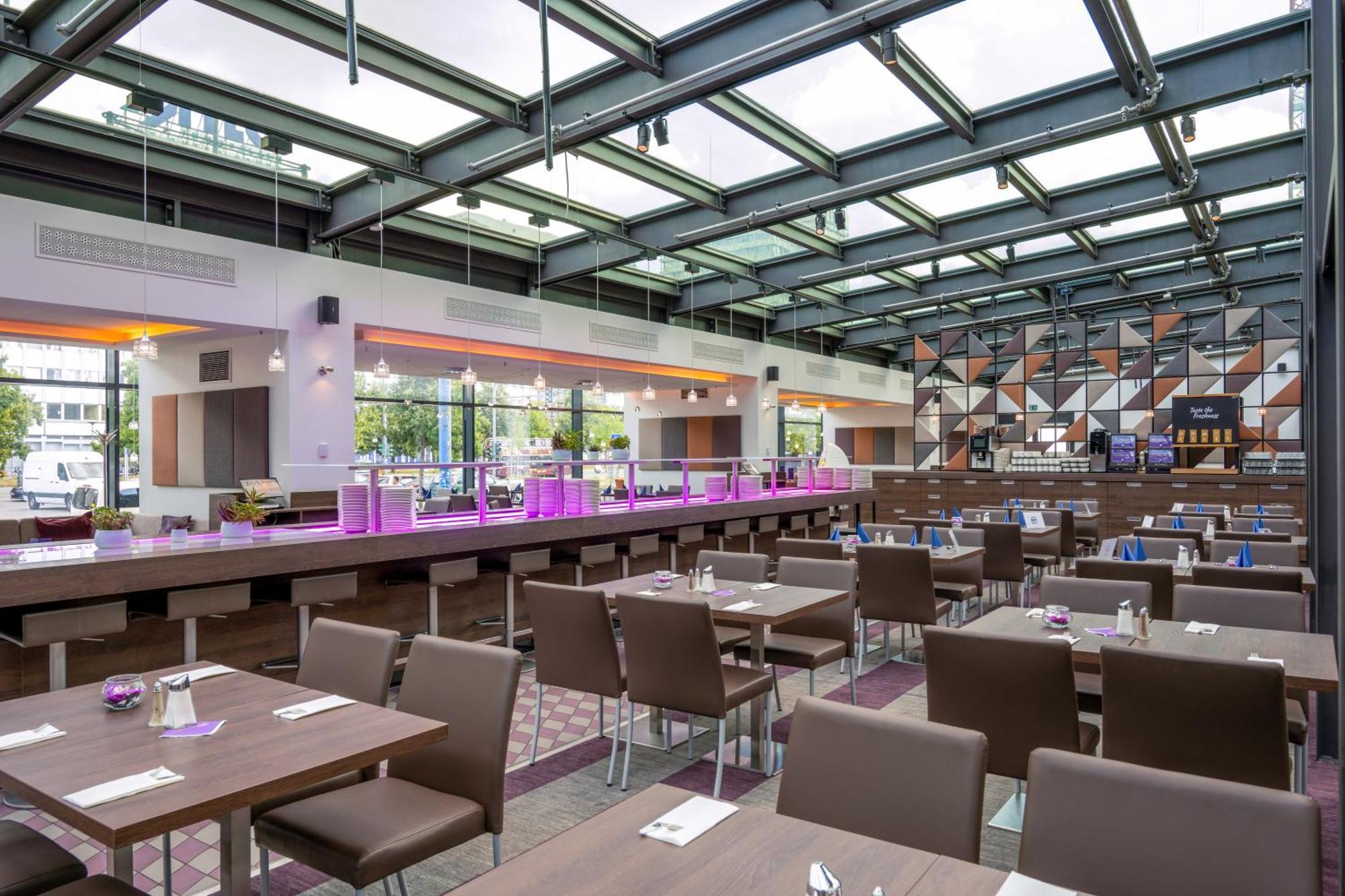 Park Inn By Radisson Berlin Alexanderplatz Esterno foto The photo shows the interior of a modern restaurant. It features a spacious and bright layout with large windows allowing natural light to flow in. The ceiling is made of glass panels, enhancing the open atmosphere. 

The dining area has several dark