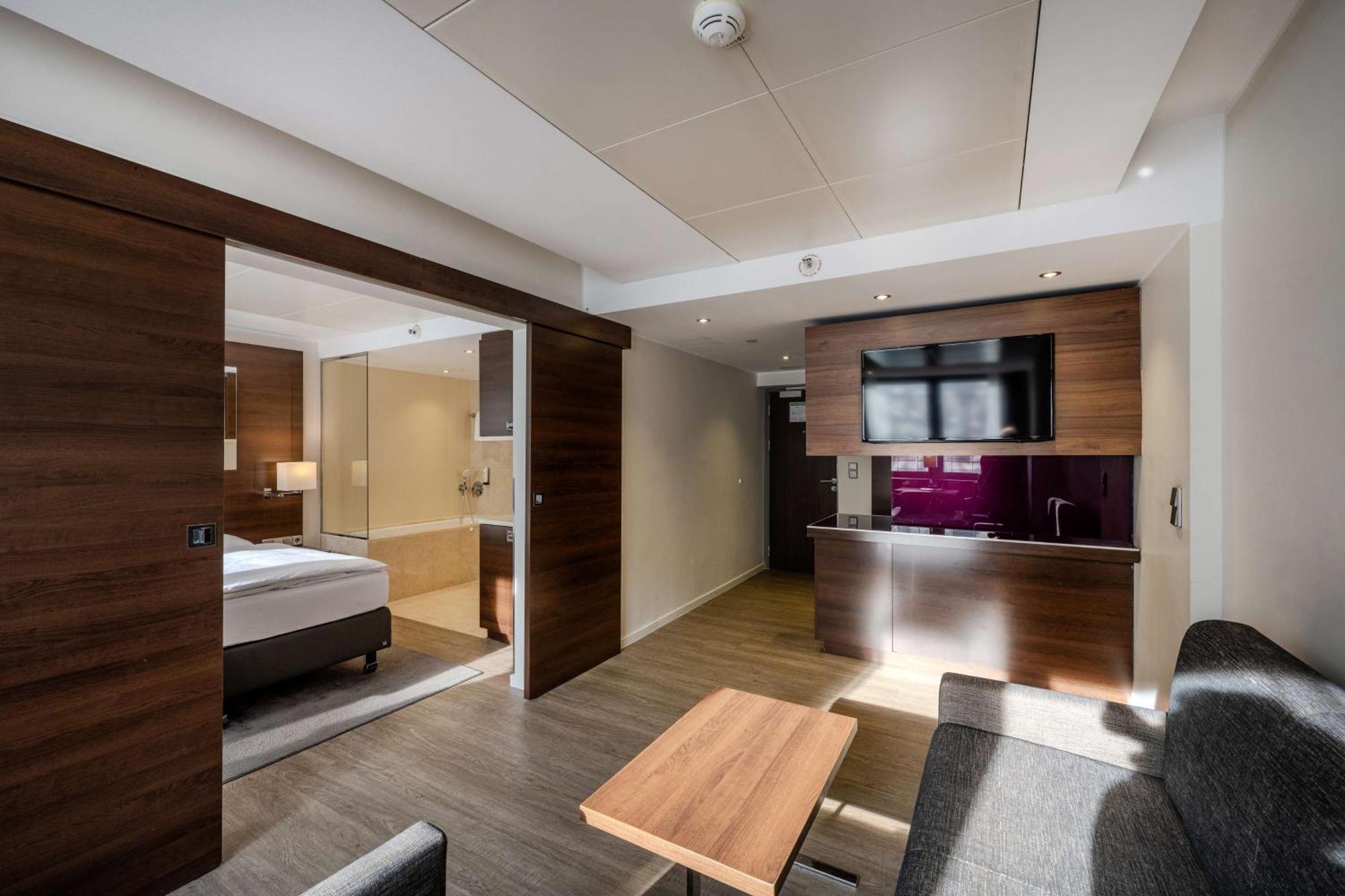 Park Inn By Radisson Berlin Alexanderplatz Esterno foto The photo shows a modern hotel room with a well-designed layout. On one side, there is a bedroom area featuring a bed and light-colored bedding. The room is separated by sliding wooden doors. The living area includes a comfortable gray couch and a wo