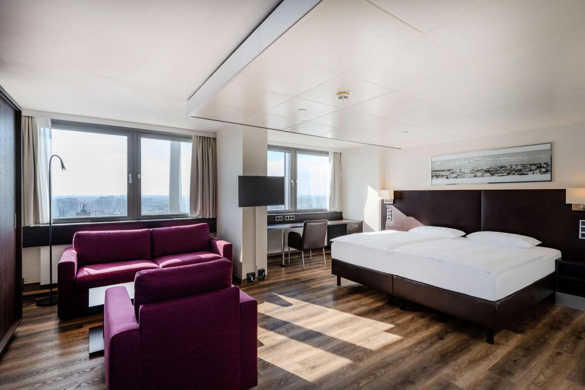 Park Inn By Radisson Berlin Alexanderplatz Esterno foto The photo shows a modern hotel room featuring a large bed with white bedding. There's a cozy seating area with a purple sofa and armchair. A desk and chair are positioned near a window, which provides natural light and offers a view of the outside. T