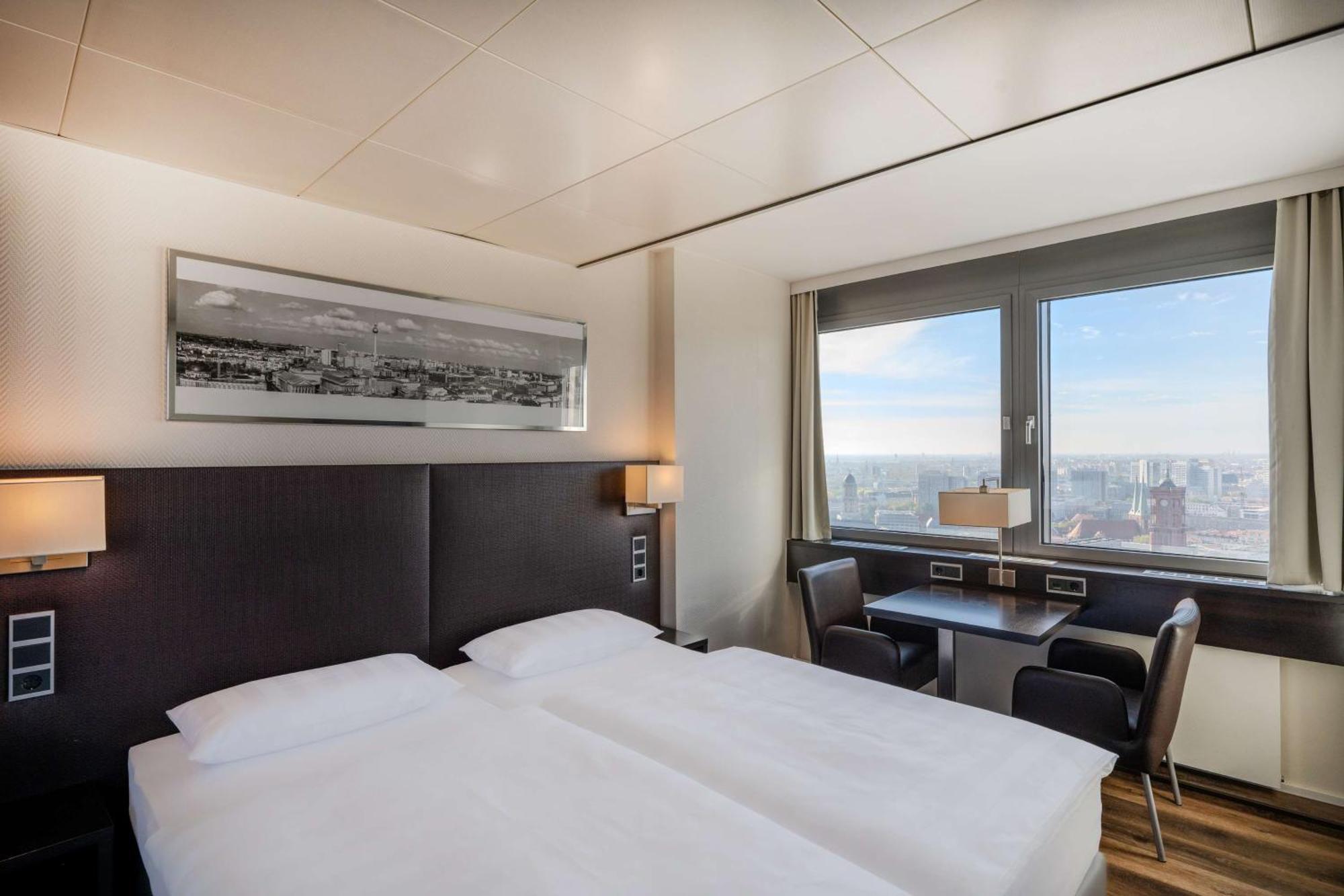 Park Inn By Radisson Berlin Alexanderplatz Esterno foto The photo shows a modern hotel room featuring two single beds with white linens and dark headboards. There is a large window that provides natural light and a view of the cityscape outside. The room is equipped with a small table and two black chairs